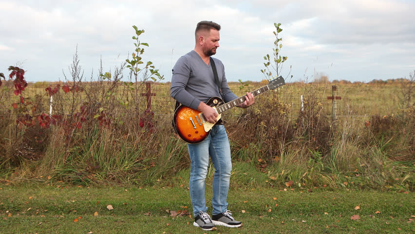 50 years old man with dark hair in a gray shirt standing on the grass having joy while playing bass guitar, musical instrument in park. Male guitarist in blue jeans resting and enjoying calm atmospher