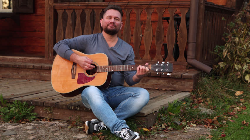 50 years old man with dark hair in a gray shirt sitting on the wood terrace while playing guitar, musical instrument in park. Male guitarist in blue jeans resting and enjoying sunset light