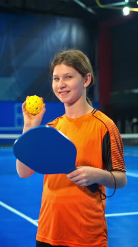A girl posing after playing pickleball on a blue, inside court. Vertical