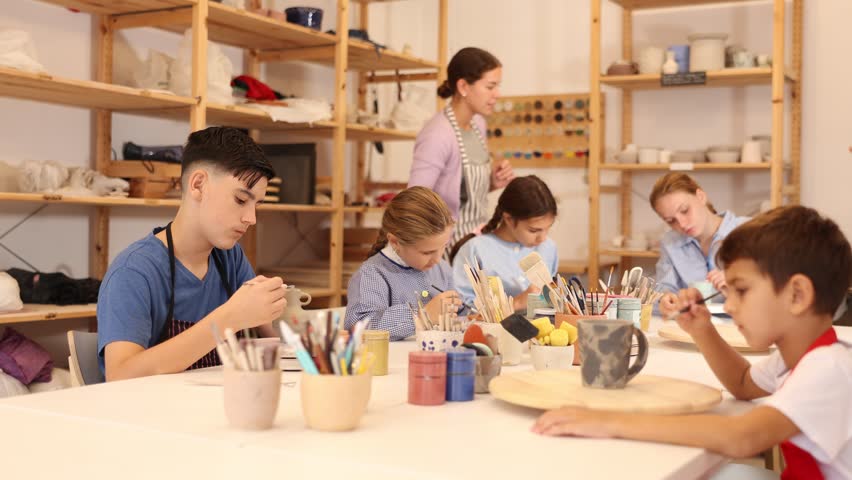 Children and teenagers sit at a table and paint ceramics at the workshop. Teacher shows how to paint a clay product after firing