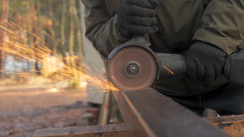 A person wearing gloves operates a cutting tool on a metal piece at an outdoor workshop, creating sparks that illuminate the surrounding forest area in the afternoon light.