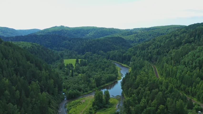 Drone camera moving forward reveals a stunning green forest alongside a river under a cloudy sky, showcasing natural beauty of Ural mountains
