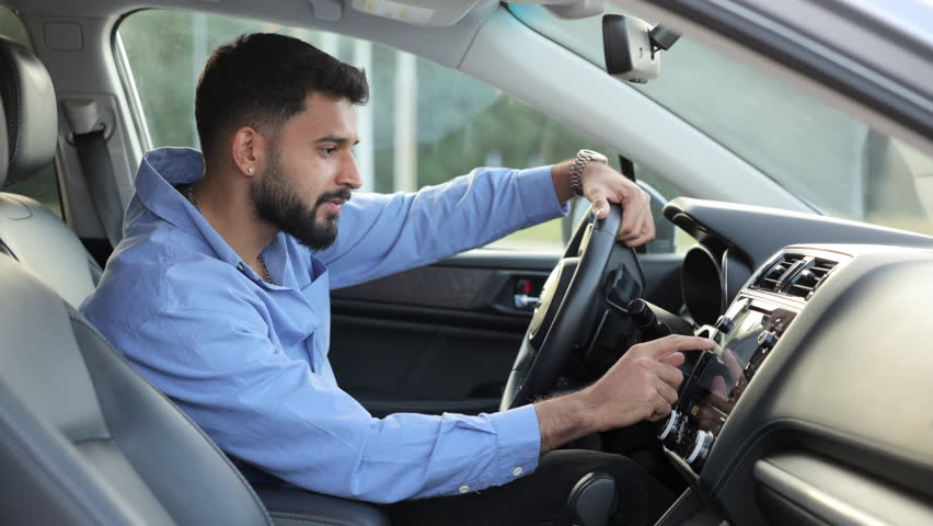 Happy owner of new car exploring functions control panel inside car. Side view of bearded male driver operating control on dashboard, going to drive car. Concept of technologies, car control.