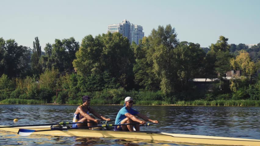 Rowing team training. Side view of 2 young caucasian male rowers, during a rowing practice, athlete sitting in a boat in the river Dnipro, rows through a calm water sunny day, autumn. 4k footage