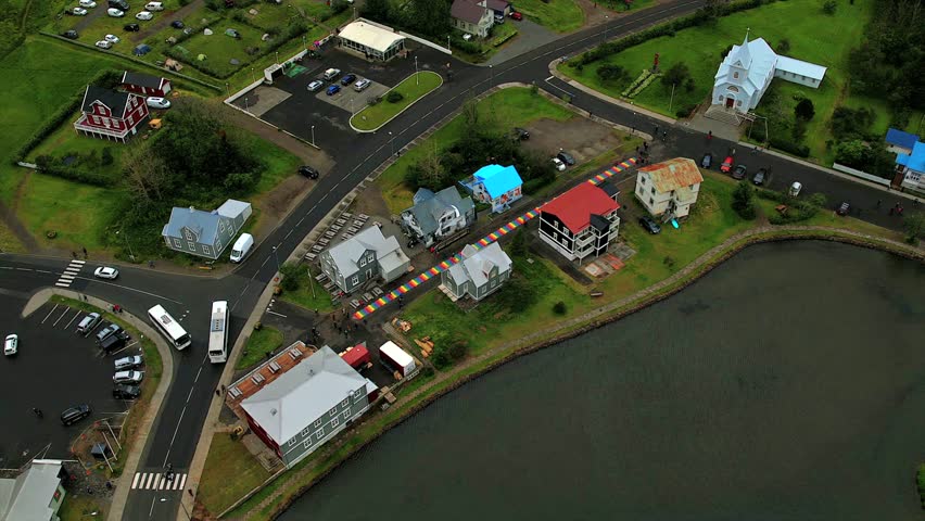 High-angle aerial view of Rainbow Street leading to the Blue Church in Seyðisfjörður, Iceland. The vibrant houses, serene waterfront, and charming streets showcase this picturesque Nordic village.