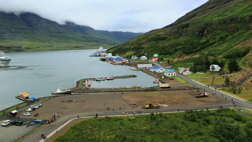 Aerial view of the harbor and colorful village of Seyðisfjörður, Iceland, with a cruise ship docked in the fjord. Surrounded by dramatic green mountains and tranquil waters.