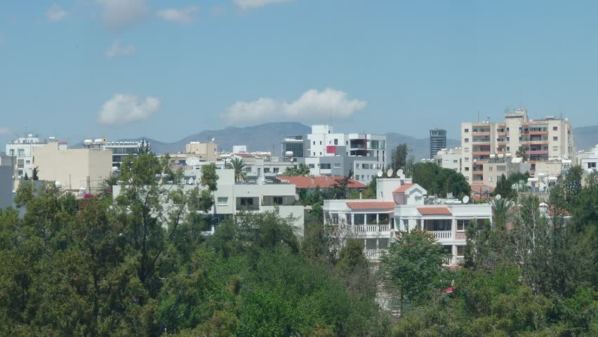 A vibrant cityscape features a mix of modern buildings and greenery under a clear blue sky, with mountains visible in the background. The scene captures urban life.
