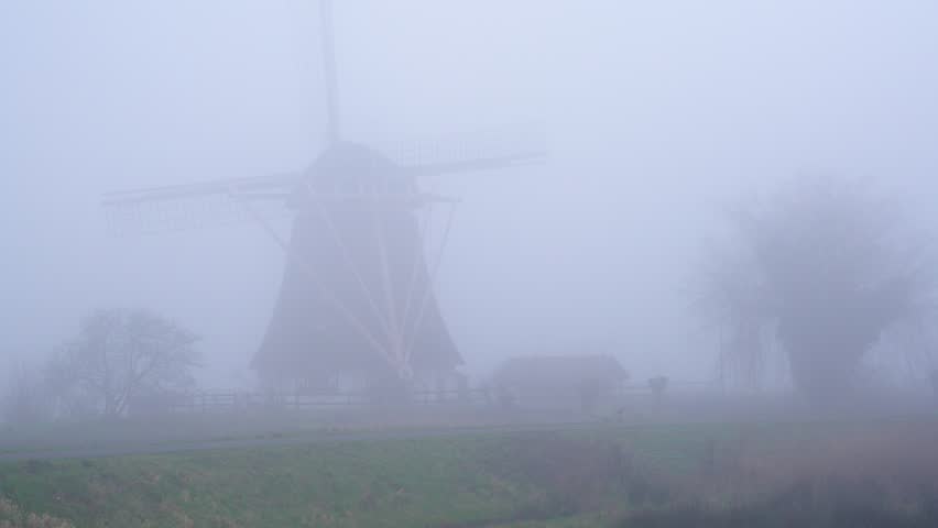 Cycling in The Netherlands in Misty Weather With Low Visibility, Cyclist passes a traditional dutch windmill in a dense fog