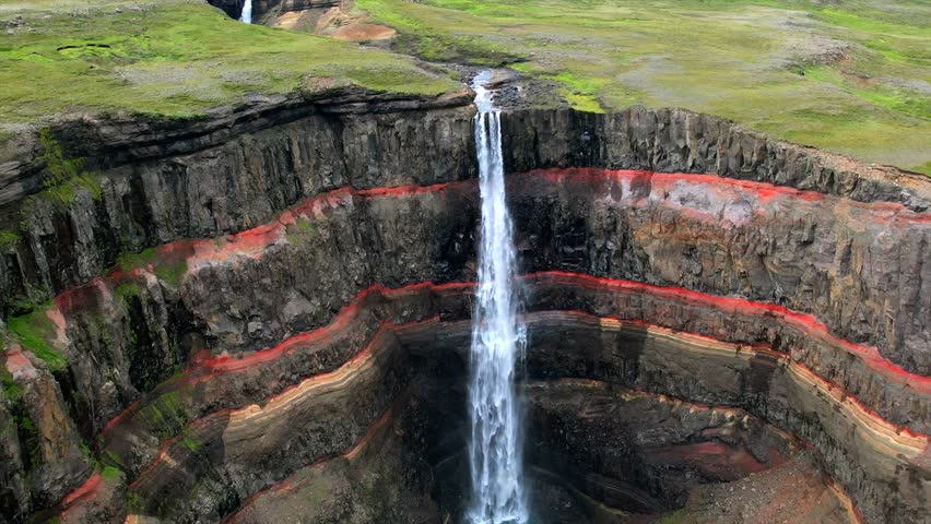 Stunning drone footage of Hengifoss Waterfall plunging down vibrant red rock layers into a deep canyon in East Iceland. Ideal for travel, nature, and cinematic projects.