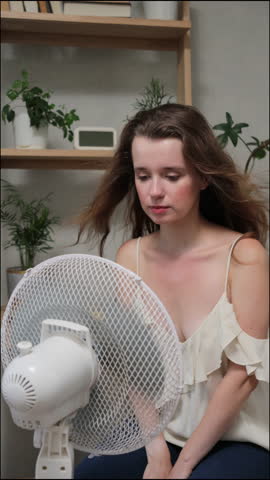 Caucasian woman is suffering from summer heat at home and is sitting in front of fan blown by wind. female housewife holds her hair in her hands for cooling. air conditioning at home