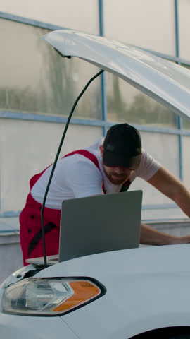 Man in uniform repairs car engine and looks at laptop