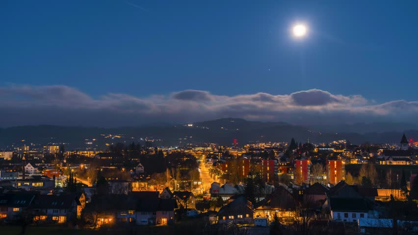 Time lapse full moon over a city with lights and car tracks. The clouds move on the horizon and the lights illuminate the city. There are small mountains in the background.
