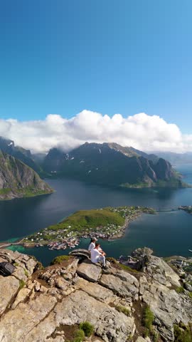 Discover the majestic beauty of Norways landscapes as a person enjoys the captivating views from a rocky summit. The tranquil waters and towering mountains, Reinebringen hike Lofoten Norway