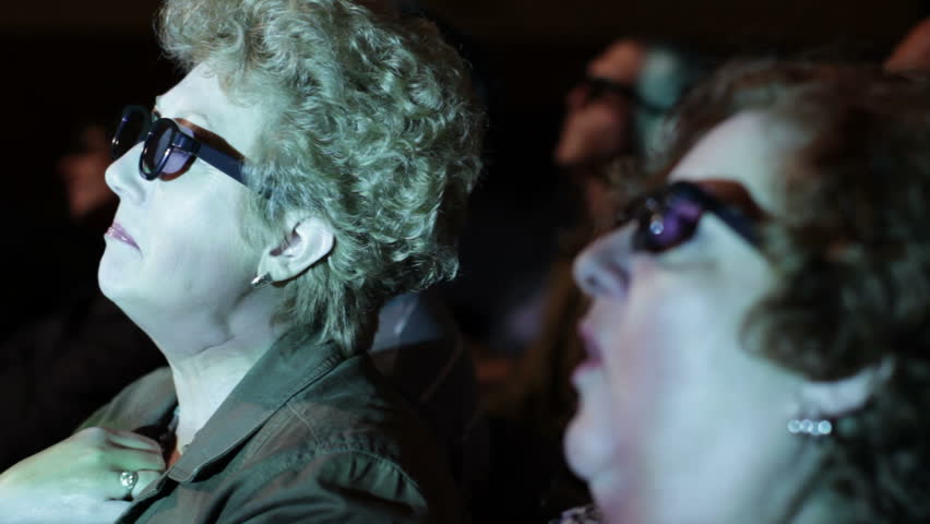 Mature woman watching a movie and wearing 3D glasses. Focus on her with a small
