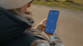 Young woman outdoors, standing on street, holding smartphone with blue screen for mockup or template, wearing winter clothes and glasses - Powered by Shutterstock - Get 15% off with code: PIKWIZARD15