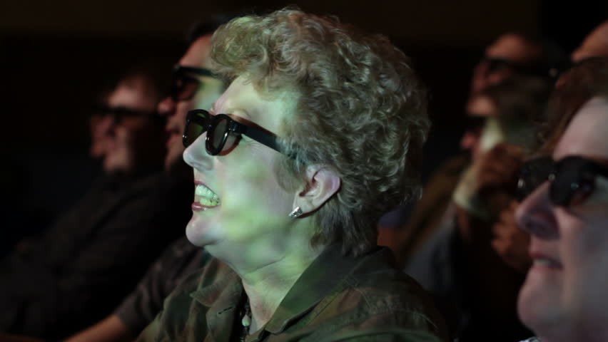 Mature woman applauds as she watches a 3D movie. Focus on her with a small dolly