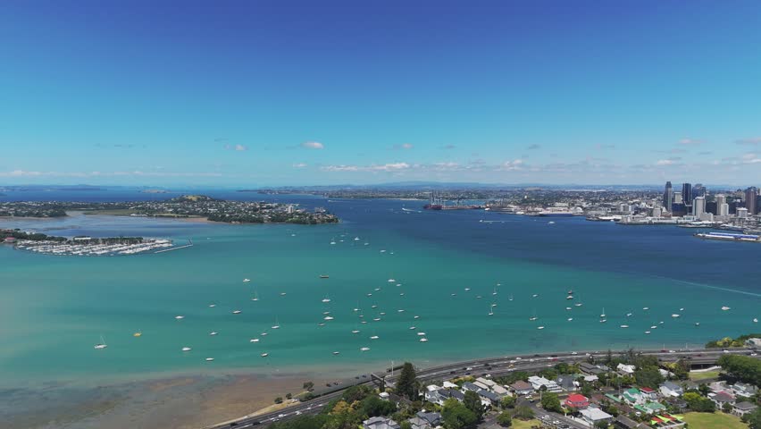 Suburb neighborhood of Auckland with traffic on coastal highway and parking boats on Shoal Bay. Auckland Skyline with tower in distance. Aerial orbit wide shot. Summer day in New Zealand.