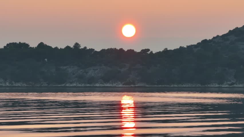 This video captures a stunning sunset over a calm sea near Sibenik, Croatia, with vibrant orange and pink hues reflecting on the tranquil water, creating a peaceful and mesmerizing scene of nature.