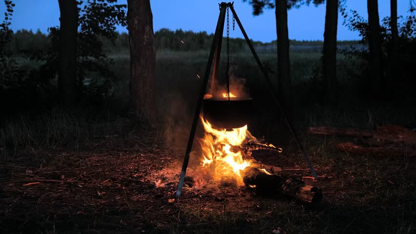 A large cooking pot hangs above a crackling campfire in a serene forest setting during twilight. Flames flicker against the darkening sky as the meal cooks.
