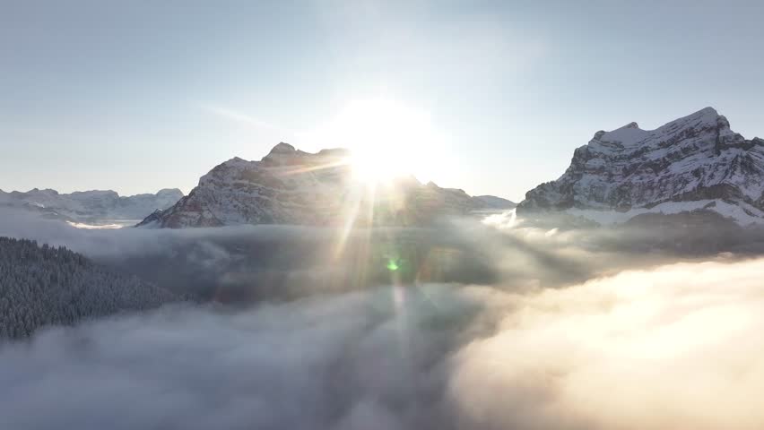 Aerial view of the Rautispitz and Glärnisch peaks in Glarus, Switzerland, surrounded by dense clouds and illuminated by a radiant sunrise. Stunning alpine scenery.