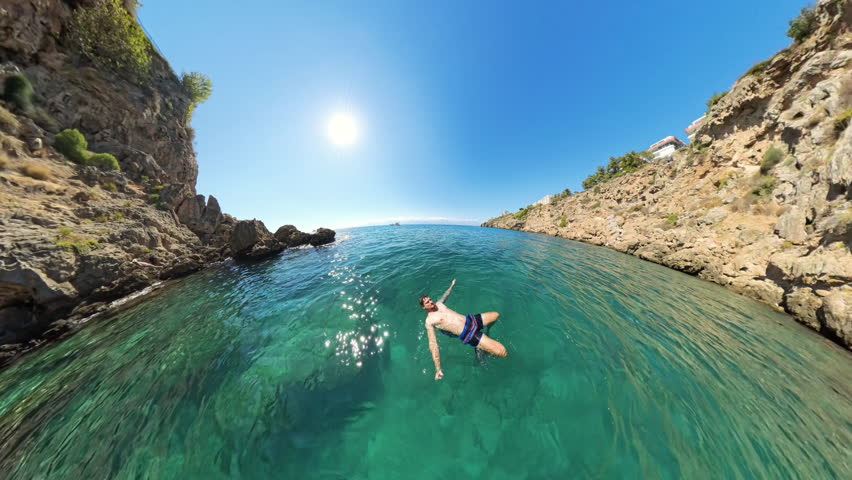 A man floating on his back in a serene, clear blue cove, framed by rocky cliffs and under a bright sunny sky, capturing the peacefulness of the moment.