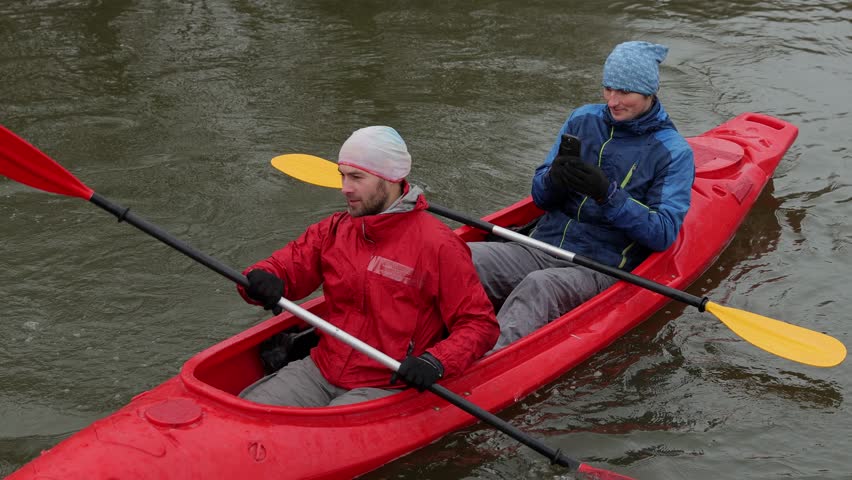 Couple paddling in a red kayak. One with a phone in his hands, the other with an oar.