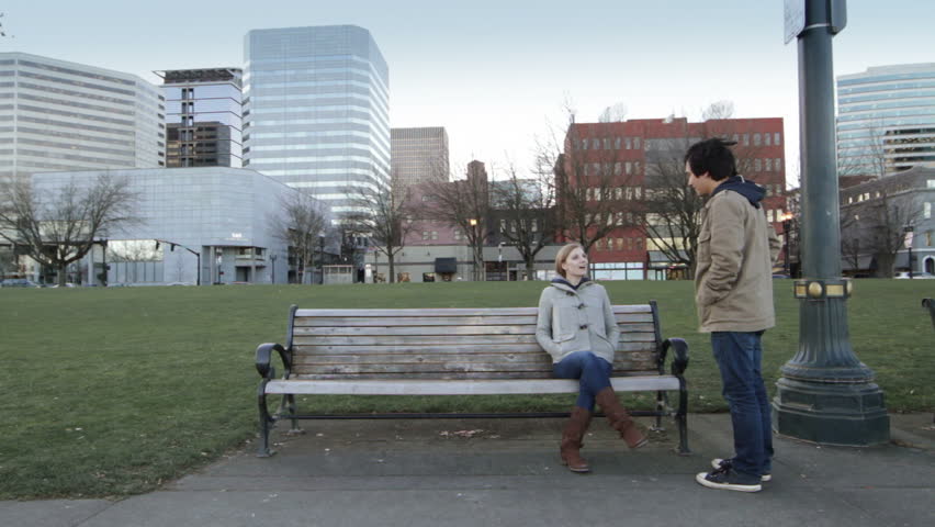 Young woman sits on park bench, young man walks past and then starts talking to