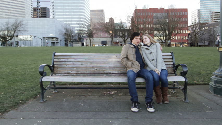 Young couple talk and embrace on a park bench. Wide shot with the city of