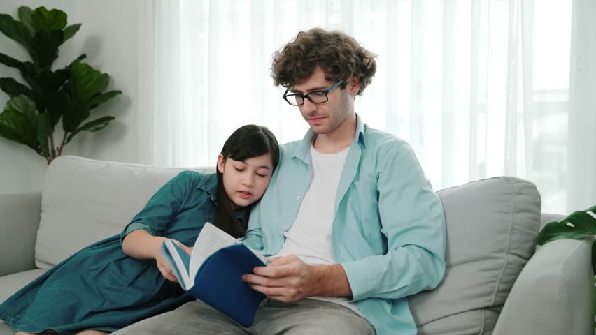 Caucasian father and cute daughter reading story book together while sitting at sofa. Happy dad telling story to cute american child to imagination development while girl pointing at book. Pedagogy.