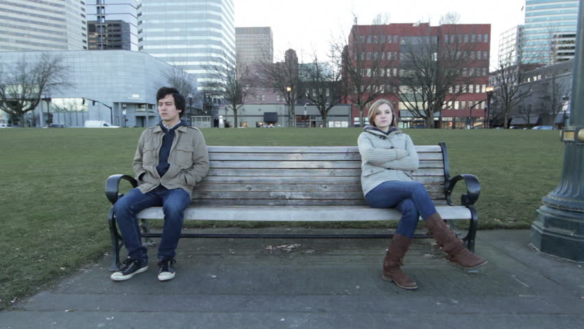 Young couple sit start at opposite sides of a park bench, frustrated with each