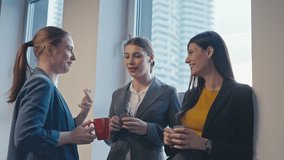 Three professional women in business attire are engaged in a casual conversation while holding coffee mugs in a modern office setting. Concept of workplace communication - Powered by Shutterstock - Get 15% off with code: PIKWIZARD15
