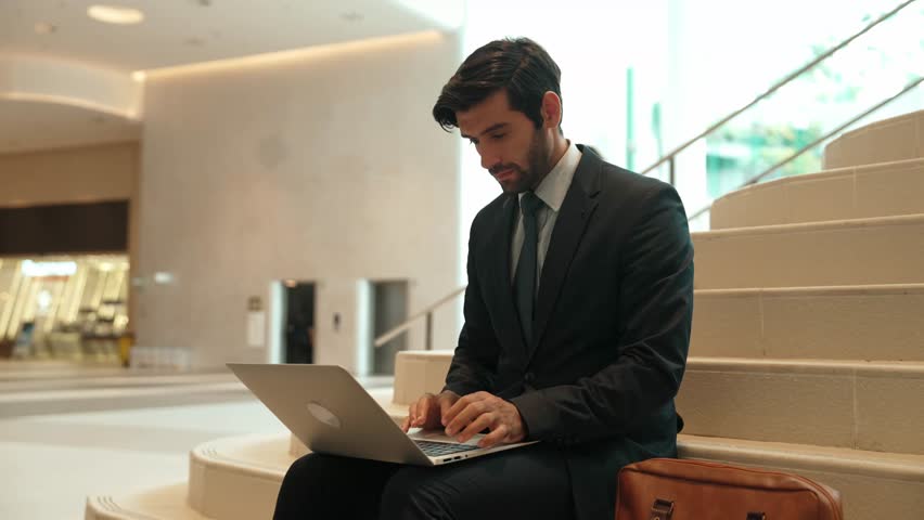 Skilled investor working or planing strategy by using laptop at stair. Business man wearing suit while working and sitting at stairs. Smart manager finish working and going to another place. Exultant.