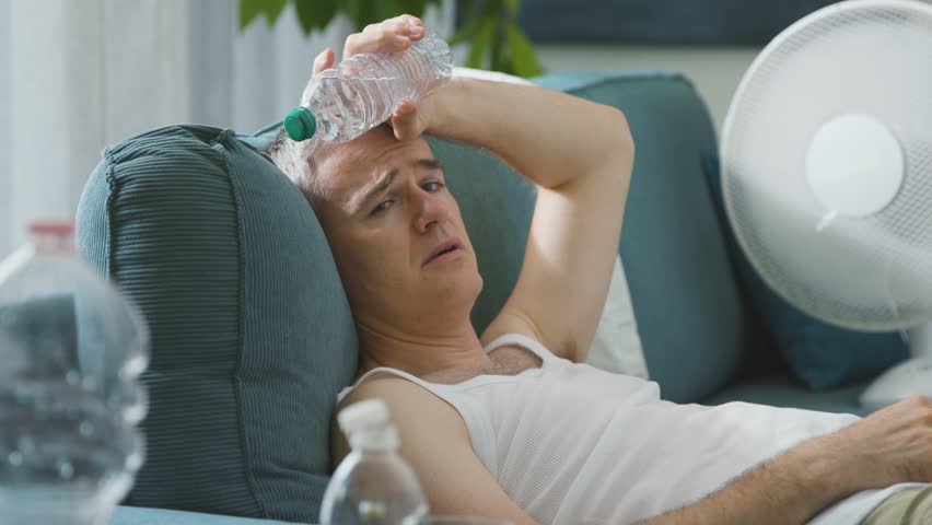 Exhausted man sitting on the couch at home during the summer heatwave, he is suffering from the heat