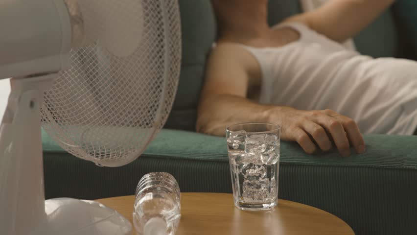 Man suffering from the heat at home, he is sitting on the couch next to an electric fan and holding a fresh water bottle