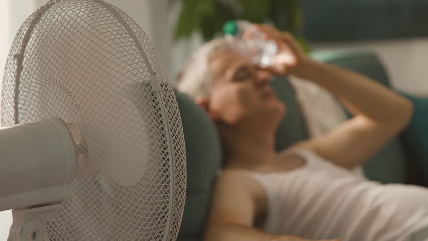 Man suffering from the heat at home, he is sitting on the couch next to an electric fan and holding a fresh water bottle