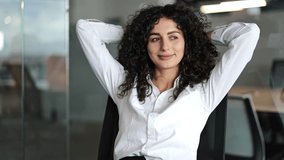 Confident businesswoman with curly hair sits relaxed in a modern office. She smiles and leans back, enjoying a moment of peace and quiet. - Powered by Shutterstock - Get 15% off with code: PIKWIZARD15