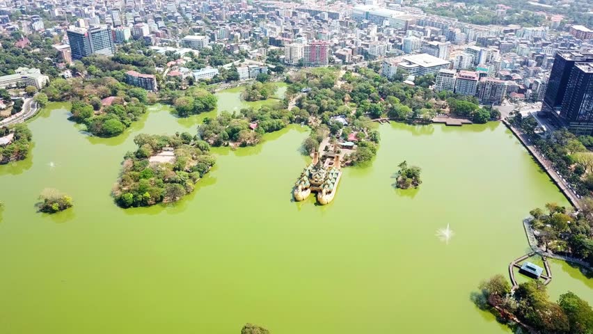 Aerial view of Kandawgyi Lake and Karaweik Palace in Yangon, Myanmar, showcasing the vibrant green water, lush greenery of the park, and the bustling cityscape in the background on a sunny day