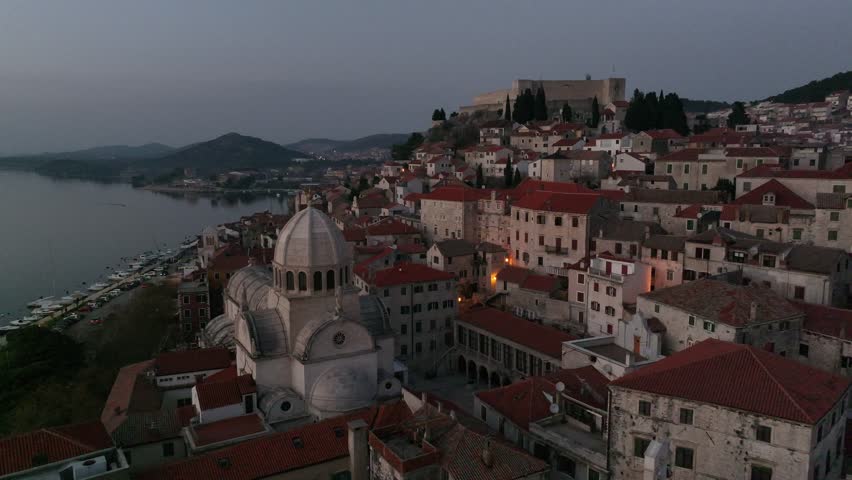 A serene aerial view of Sibenik, Croatia, showcasing the intricate dome of St. James Cathedral, the surrounding stone houses with terracotta roofs, the tranquil hillside and waterfront during sunset.