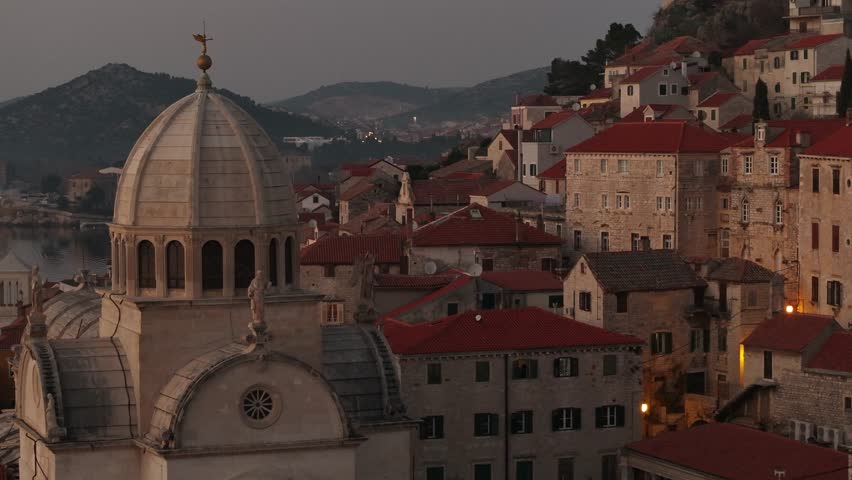 A breathtaking view of Sibenik, Croatia, highlighting St. James Cathedral, a UNESCO World Heritage Site, surrounded by stone buildings, terracotta rooftops, and scenic hills under the sunset.