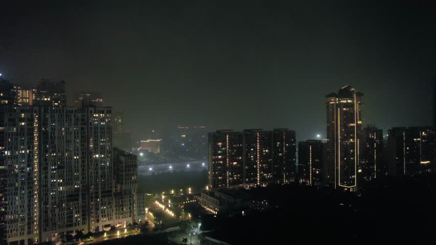 Aerial drone night shot flying towards lit skyscrapers, homes, houses, office buildings covered in fog haze showing the cityscape and polluted winters of delhi, noida, gurgaon, bangalore