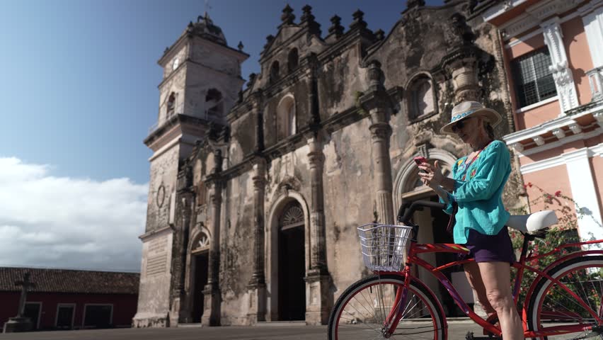 A mature woman enjoys biking outside La Merced church in Granada, Nicaragua. She captures her experience on her phone, soaking in the vibrant surroundings.