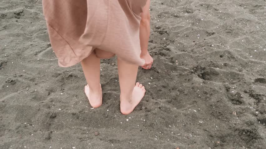 A woman enjoys the sandy beach and plays with the grains. A closeup of a white woman in a beige dress playing with her hand in the sand on a warm beach during the light breeze.