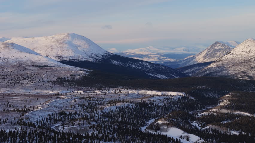 A cinematic aerial view of Mountain Landscape near Fish Lake at sunset. Golden hour light enhances the frosty Yukon wilderness, showcasing a pristine snow-covered winter scene.