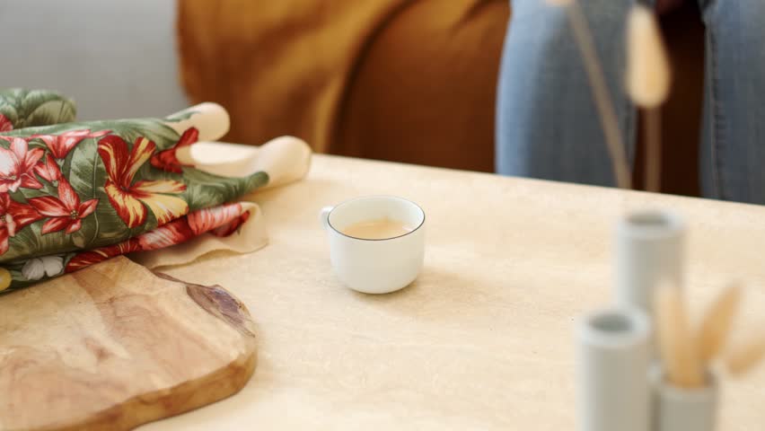 Hand picks up a small white coffee cup and sets it down on a table with floral fabric