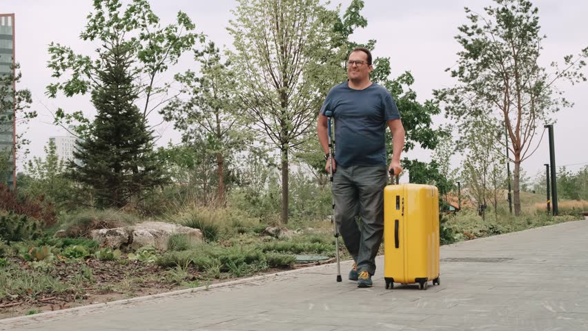 An adult caucasian man with glasses barely hobbles on one leg with crutches in park with large yellow tourist suitcase. Disabled male traveler concept. Middle-aged tourist walking in the city center