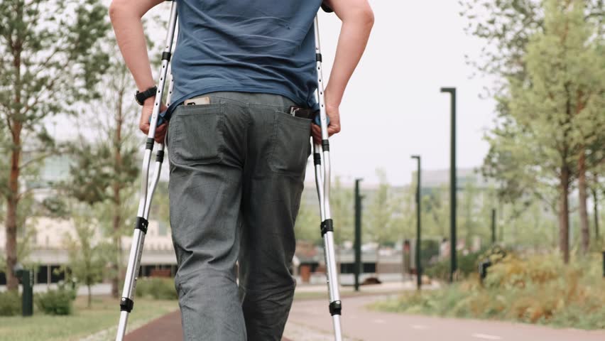 Close-up of man barely hobbles on one leg with crutches. Male in sneakers and gray pants walks slowly down the street, leaning on two crutches. Disabled person walking in park during day. Rear view