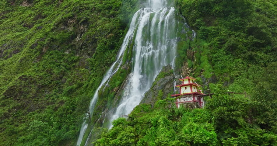 Drone gliding toward the stunning Narchyang Waterfall, with the picturesque Bhumethan Temple visible in the foreground, set against the lush green landscape of Nepal