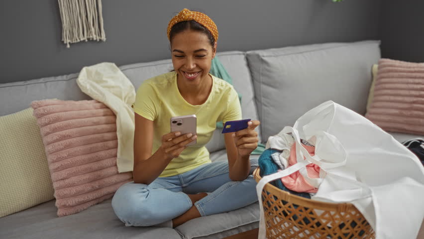 Young woman sitting in living room shopping online with mobile phone and credit card surrounded by folded laundry