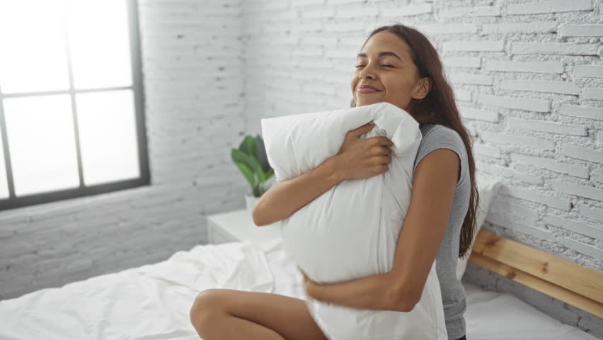Young woman in bedroom hugging pillow with a smile, sitting on bed, enjoying the peaceful morning light through the window, in a minimalist home interior