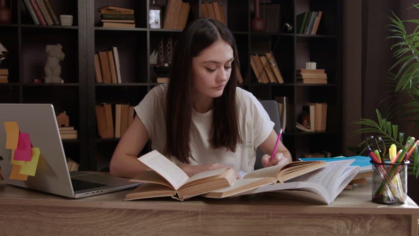 A cheerful young female student is browsing through an old book in the library and taking notes for school. A classic library and a girl enthusiastically reading specialized books for studies